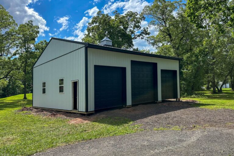 Featured Project 7 Black and white garage with three garage doors and two windows and a door on the side facing the camera set in a gravel driveway with green grass and blue sky